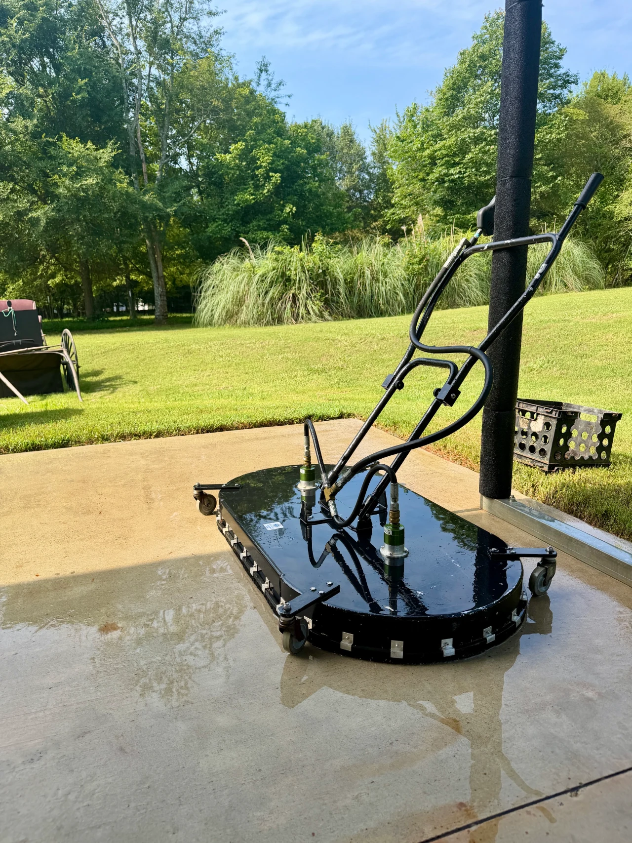 Large rotary surface cleaner resting on wet concrete beneath a carport.